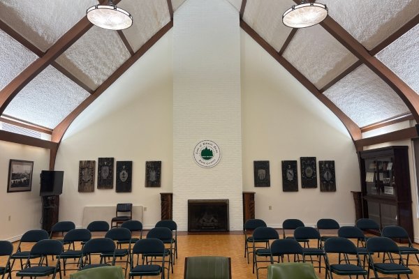 The interior of the Biltmore Forest Town Hall gathering hall — a vaulted barrel ceiling with curved wooden beams, white painted brick walls, rows of green folding chairs facing the central brick fireplace at the back wall where the Echoes of the Forest Black Walnut mantel will be installed, with the Biltmore Forest town seal mounted above