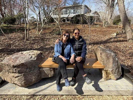 The Tea for Two bench by Mark Oliver of Foundation Woodworks installed in the Plants of Promise Garden at the North Carolina Arboretum — a two-seat live-edge bench handcrafted from Hurricane Helene–salvaged timber, sited between the Education Center and the Playing Woods, framed by spring greenery