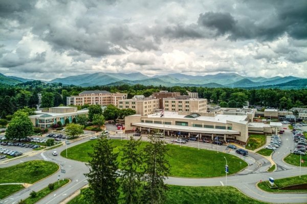 An aerial view of the Charles George VA Medical Center campus in Asheville, North Carolina — a multi-building hospital complex set among rolling green hills, with the Blue Ridge Mountains rising in the misty distance under a sky of dramatic clouds, the future site of the July 4 2026 Echoes of the Forest veterans' memorial bench