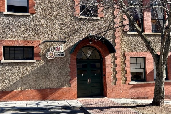 The brick and stucco facade of the historic YMI Cultural Center in downtown Asheville, North Carolina — the round YMI Cultural Center logo mounted beside the green double-door entrance, a bare tree casting shadows across the building, in the heart of The Block, Asheville's historic African American district