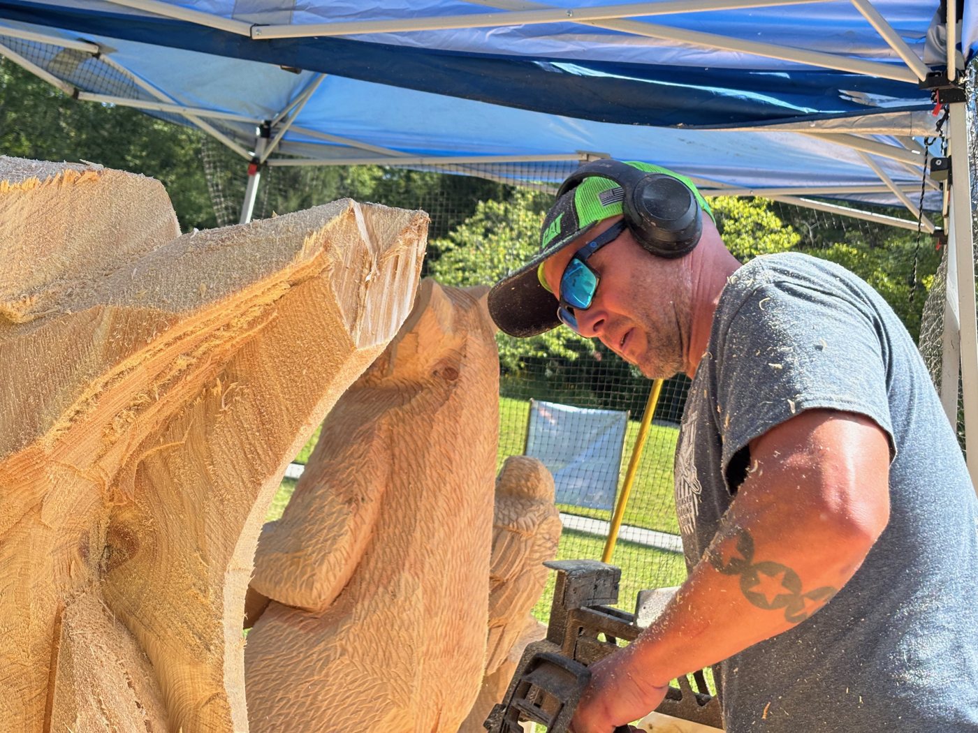 Mike Ayers in ear protection and sunglasses, carving a wooden animal with a power tool under a tent at the Cradle of Forestry