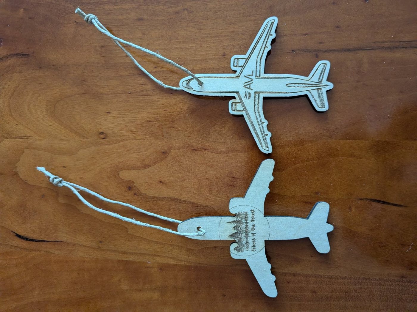 Two laser-cut wooden airplane ornaments on a wood table, one engraved with the AVL logo and one with the Echoes of the Forest logo, each on a twine hanging loop