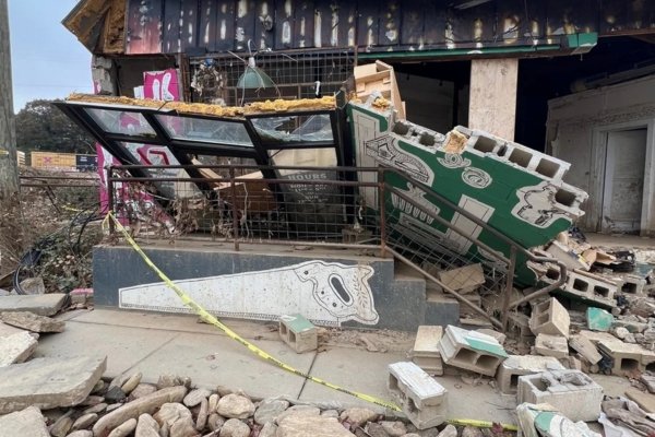 The exterior of Foundation Woodworks gallery in Asheville's River Arts District after Hurricane Helene receded — the entrance collapsed, broken windows lying flat on the porch, cinder block walls toppled, debris strewn across the front, yellow caution tape across the property, and a saw graphic still visible on the front planter from the gallery's pre-flood signage