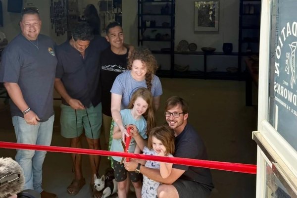 Mark Oliver kneeling with his family beneath the new Foundation Woodworks tent gallery banner in Asheville's River Arts District, two young children holding the red ribbon and scissors at the official reopening ceremony — staff and supporters smiling alongside as the gallery officially reopens after the Hurricane Helene flood