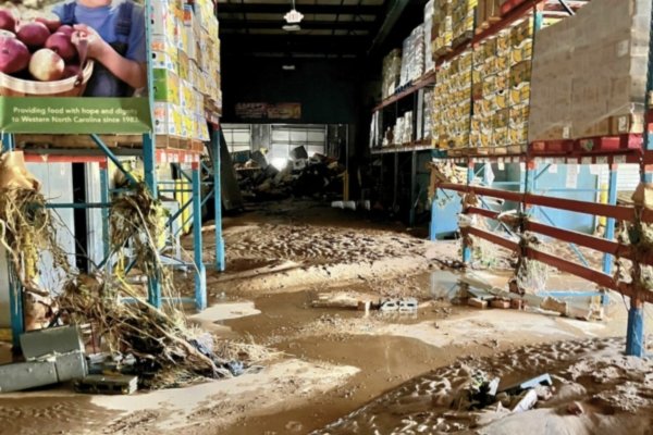 The interior of MANNA Food Bank's Asheville warehouse after Hurricane Helene's floodwaters receded — pallets of food destroyed, mud caked across the floor, and a banner visible reading 'Providing food with hope and dignity to Western North Carolina since 1983'