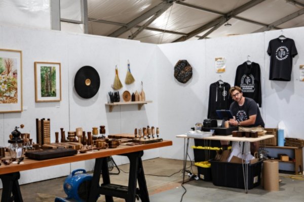 Mark Oliver, woodworker and Echoes of the Forest artisan, at the front desk of Foundation Woodworks' temporary tent gallery in Asheville's River Arts District — surrounded by his handcrafted wood pieces, framed prints, and Foundation Woodworks T-shirts displayed for sale, after the original gallery was destroyed in the Hurricane Helene flood