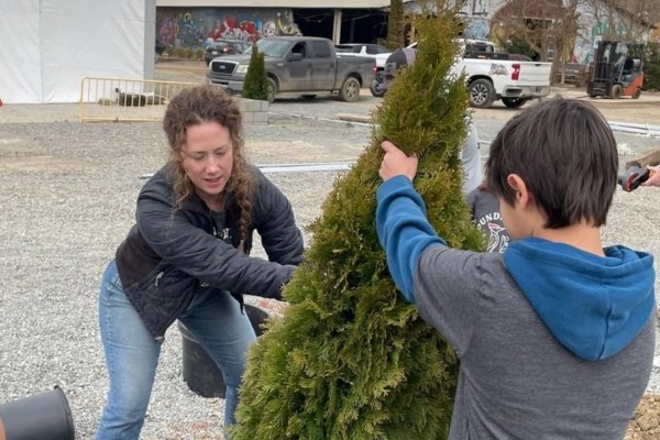A woman and a young person planting a young evergreen sapling in fresh gravel outside Foundation Woodworks' temporary tent gallery in Asheville's River Arts District — the colorful murals of the surrounding RAD buildings visible in the background, marking the literal replanting of a neighborhood that flooded under 24 feet of water during Hurricane Helene