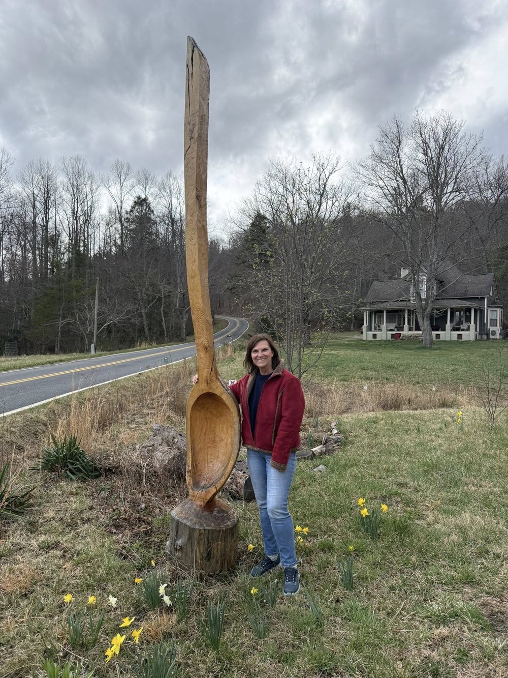 Liisa Andreassen standing beside a tall hand-carved wooden spoon sculpture outside Paul Eisenhauer's studio in Celo, NC, with daffodils blooming at the base