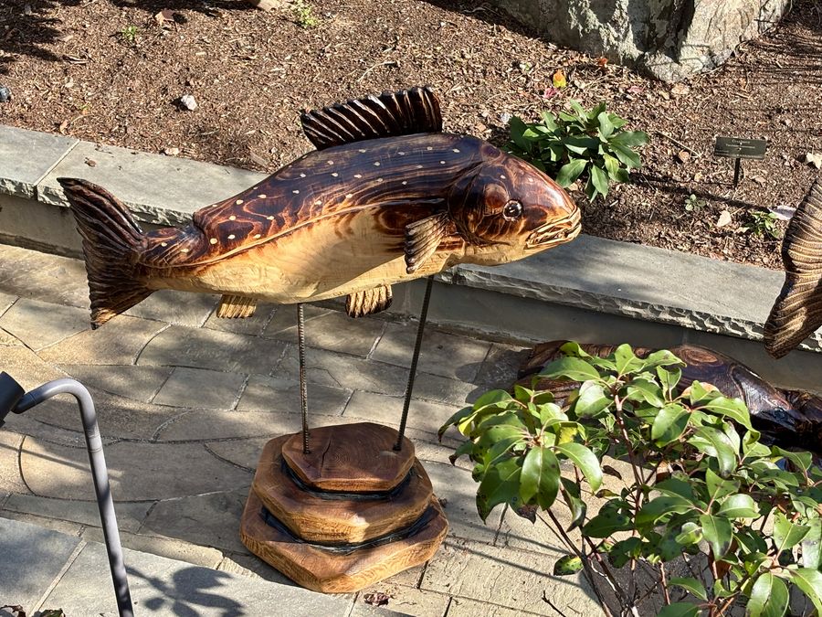 A hand-carved wooden fish sculpture mounted on a stacked wood pedestal at the North Carolina Arboretum