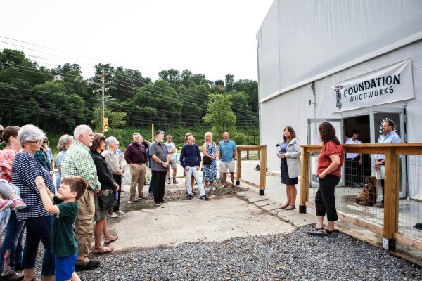 A community gathered outside Foundation Woodworks in Asheville for an Echoes of the Forest event, listening to a speaker introducing the project