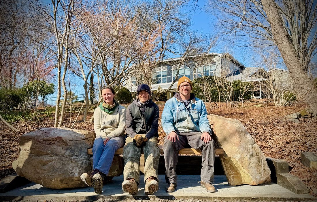 Three artisans seated together on the Tea for Two memorial bench in a sunlit winter garden at the NC Arboretum