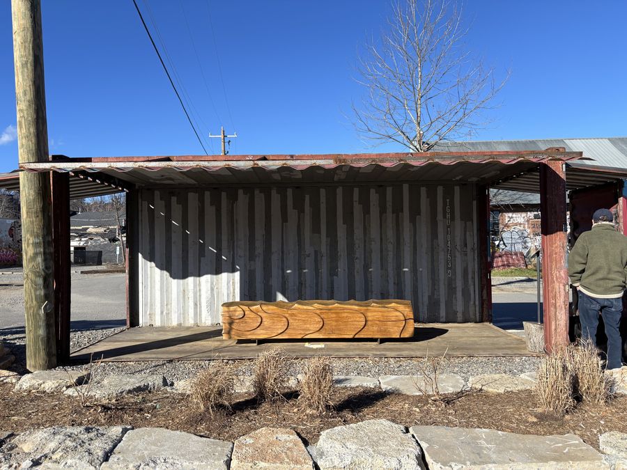 A long sculptural log bench with rippling wave-form carving, displayed inside a metal shipping container at a workshop