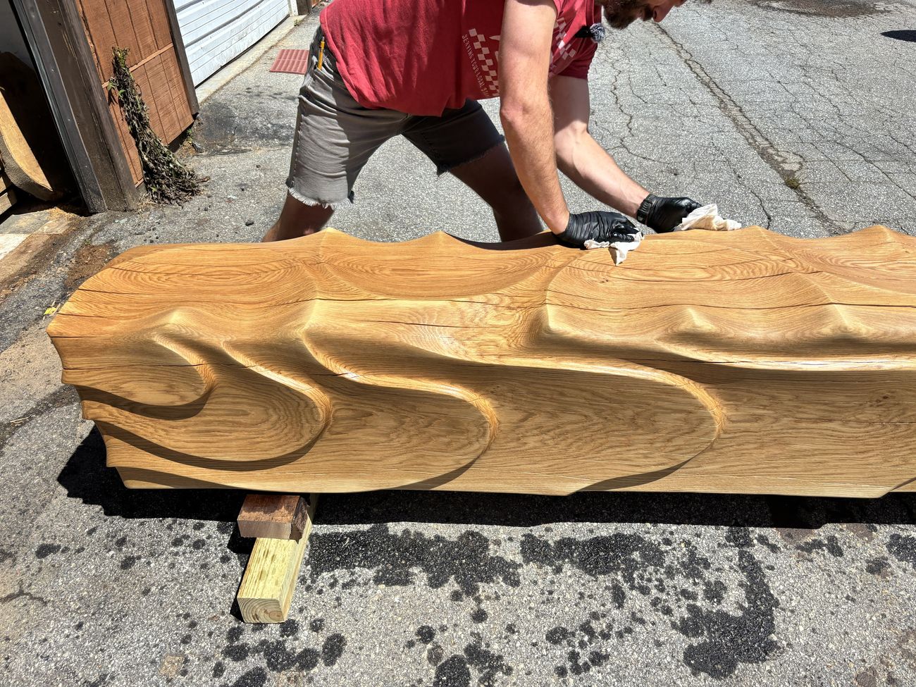 A craftsman wiping down a long, sculptural wave-form oak bench in a workshop alley, showing the bench's rippling carved surface