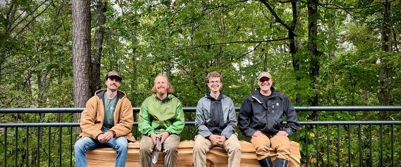 Four people seated together on the live-edge Topographical Bench, a piece of Hurricane Helene–salvaged wood by artist Chester Shuey, on a railed overlook at the North Carolina Arboretum