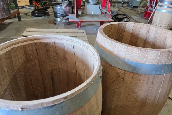 Two empty oak barrels sit on the floor of the Appalachian Cooperage workshop in Marion, North Carolina — staves freshly assembled and banded with steel hoops, raw oak grain visible inside, made from Hurricane Helene–salvaged white oak for the Echoes of the Forest x Burial Beer Co. Brown Ale collaboration