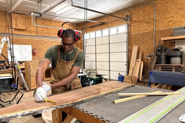 Kwadwo Som-Pimpong of Crafted Glory working in his Asheville woodshop — wearing safety glasses and over-ear hearing protection, he's bent over a long live-edge slab of richly figured hardwood, measuring and marking it with a tape measure and pencil, surrounded by his tools, lumber, and a Festool track saw