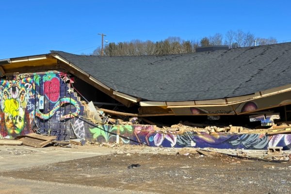 An artist studio in the River Arts District with a partially collapsed roof and graffitied murals still intact across the front facade, weeks after Hurricane Helene's floodwaters receded