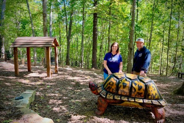Liisa Andreassen and Mike Ayers standing behind 'Shelly,' the 800-pound white pine box turtle sculpture, in the Playing Woods at the North Carolina Arboretum — Shelly's intricately carved shell rendered in shades of amber, gold, and deep brown, with a small interpretive shelter visible behind in the dappled sunlight of the surrounding forest