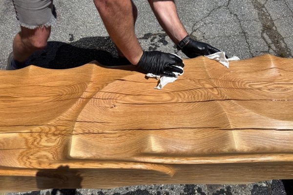 A close-up of an Echoes of the Forest woodworker's gloved hands applying finish to the carved wave hollows of a live-edge bench on a sunny driveway — the white oak grain glowing under the wipe of finish, every wave catching the light