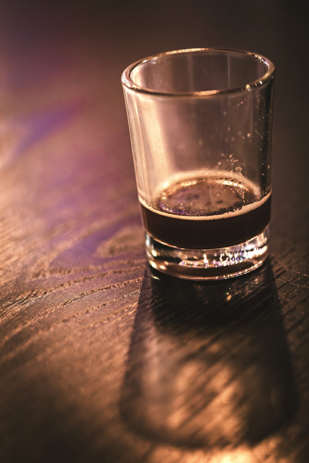 A small tasting pour of Echoes of the Forest Oak Aged Brown Ale on a wooden bar, lit warmly from above