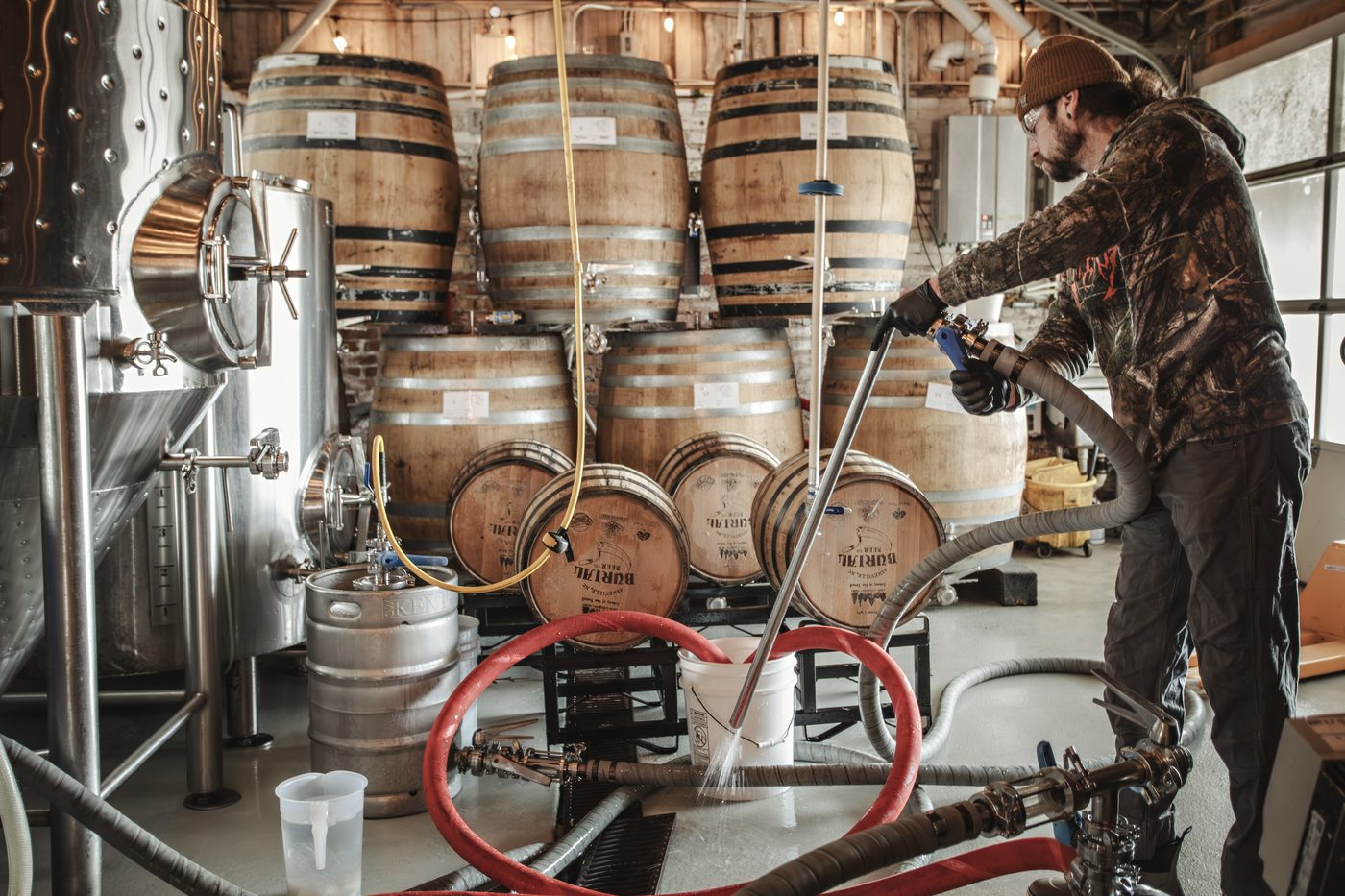 Tim Gormley cleaning brewing equipment beside a wall of Echoes of the Forest white oak barrels at Burial Beer Co.'s Forestry Camp