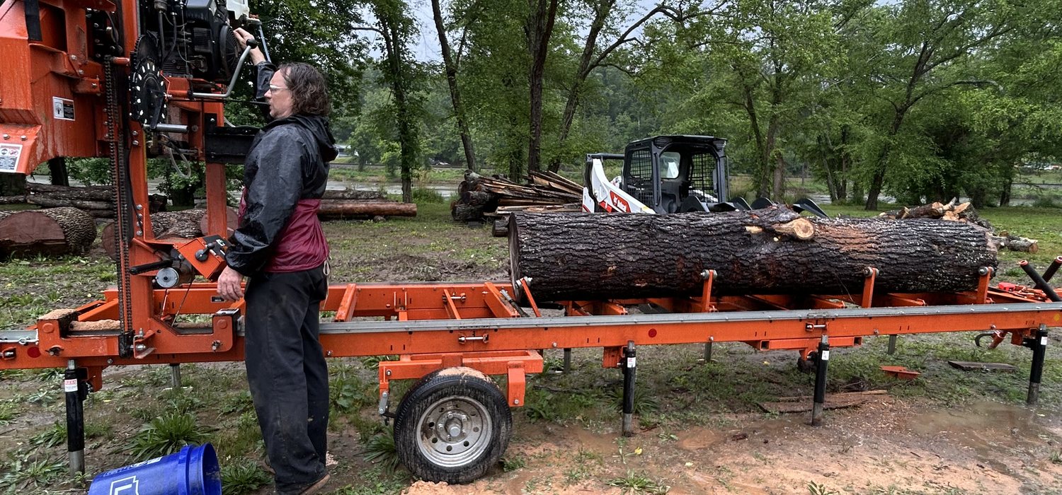 The mill operator runs a Helene-felled log through the orange portable sawmill at UNC Asheville's Community Lumber Project, with a skid-steer and salvaged timber in the background