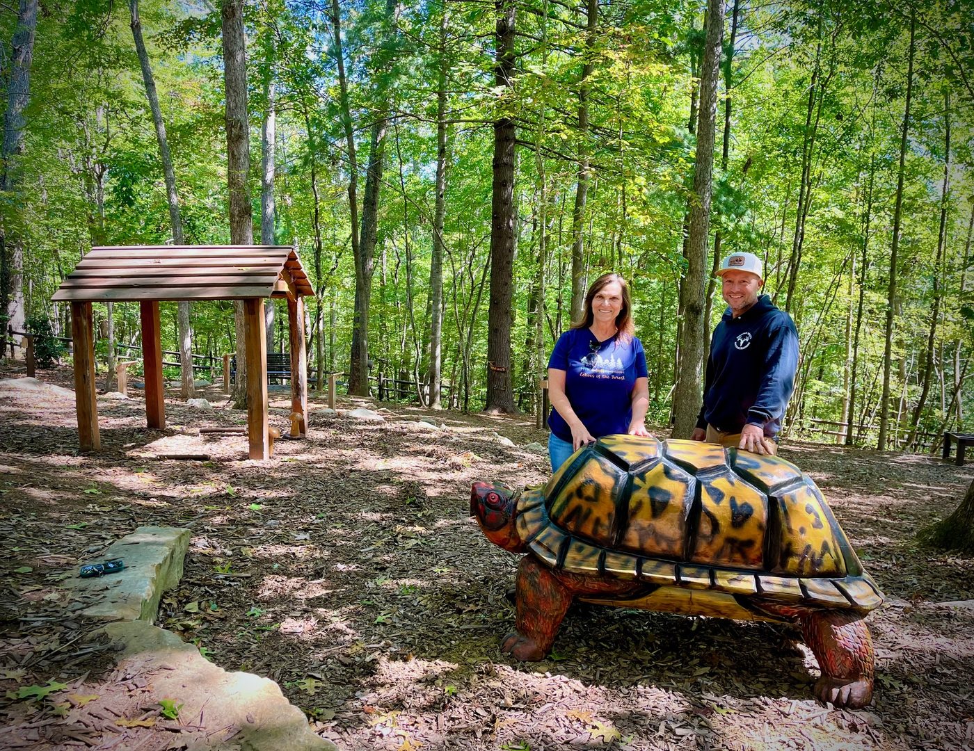 Liisa Andreassen and woodworker Mike Ayers stand beside Shelly the Box Turtle, a hand-carved wooden sculpture installed along a wooded trail at the North Carolina Arboretum